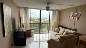 A spacious living room with a sofa, a coffee table, and a TV. There is a balcony with a view of the city. - Wonderful fully furnished 2-bedroom condo in Clearwater Beach. (Clearwater Beach)