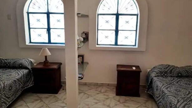 Una habitación con dos camas, una mesita con una lámpara y dos ventanas con diseño de celosía.