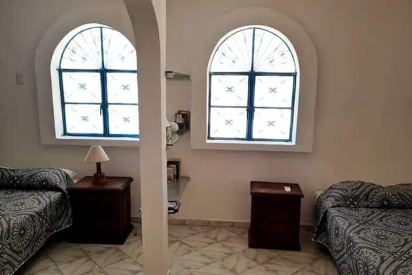 Una habitación con dos camas, una mesita con una lámpara y dos ventanas con diseño de celosía.