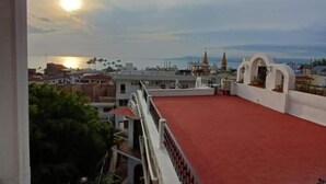 Una terraza en la azotea con piso rojo, barandillas blancas y vistas a la ciudad y al mar al atardecer.