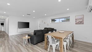 Un salon moderne avec un canapé d’angle gris, une table à manger en bois avec des chaises blanches, un téléviseur à écran plat fixé au mur et une fenêtre donnant sur l’extérieur.