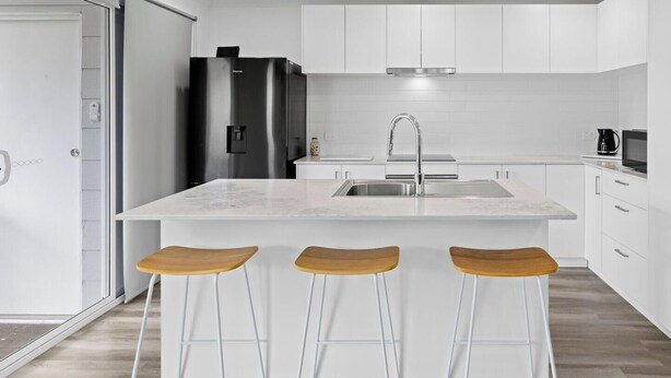 Une cuisine moderne avec des meubles blancs, un réfrigérateur en acier inoxydable, un évier et trois tabourets de bar en bois.