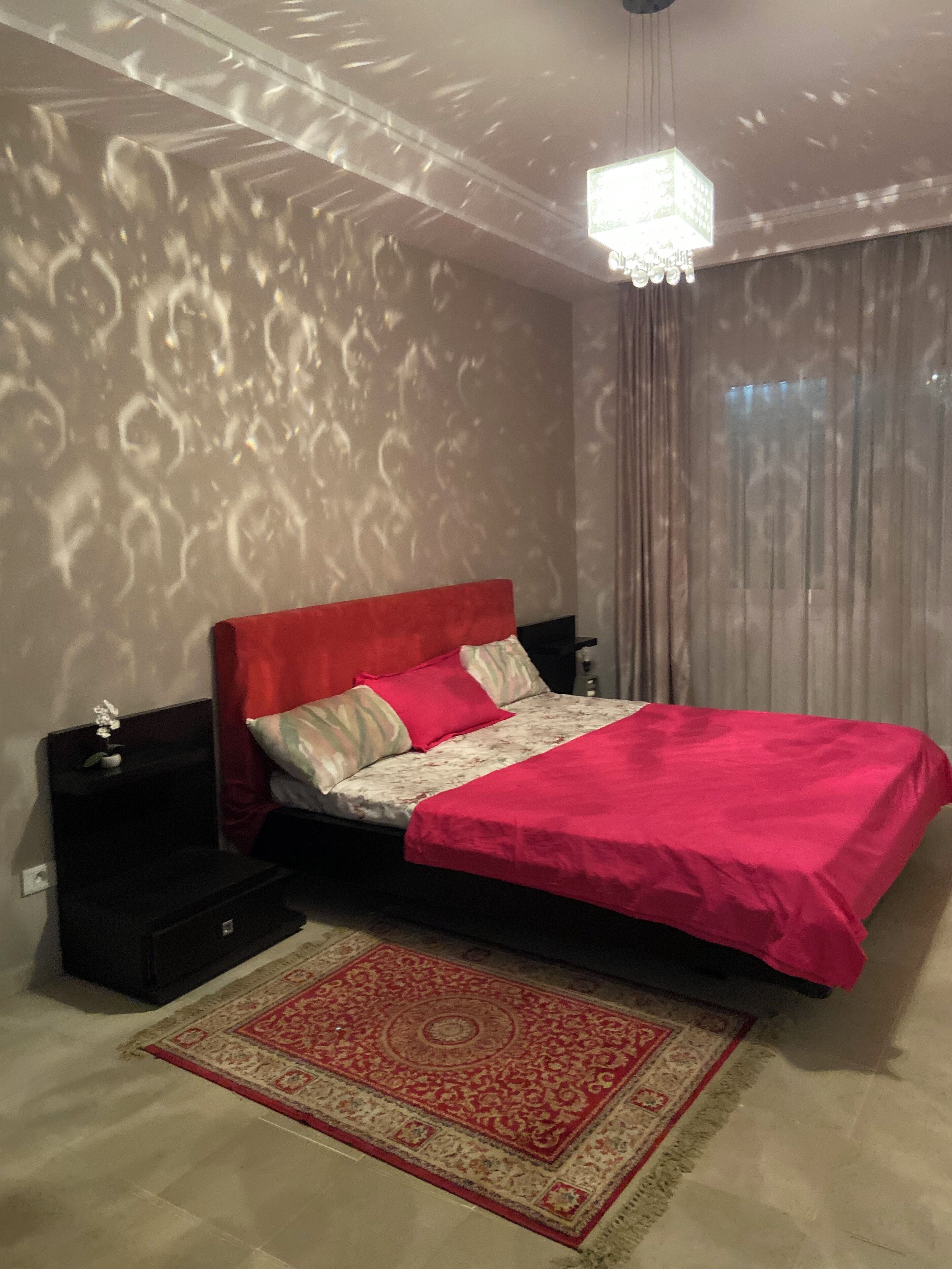A bedroom with a bed, nightstands, a chandelier, and a patterned rug.