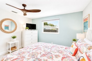 Une chambre à coucher avec un lit, un téléviseur posé sur une commode, un ventilateur de plafond, une fenêtre avec vue et un miroir décoratif.