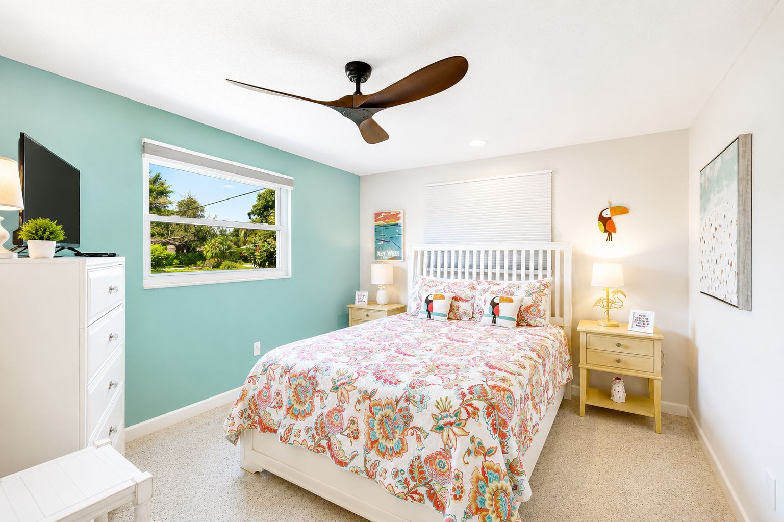 A bedroom with a bed, a ceiling fan, a window with a view, a dresser, a nightstand, and a lamp.