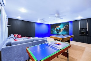 A room with a pool table, a sofa, ceiling fans, and a large wall-mounted TV.