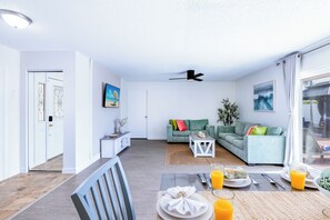 A modern living room with a dining area, a ceiling fan, and a view through a large window.