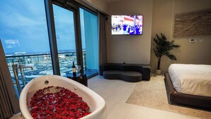 Une chambre d’hôtel moderne avec une grande baignoire remplie de pétales de roses rouges, un lit, un canapé, un téléviseur fixé au mur et un balcon offrant une vue sur la ville.
