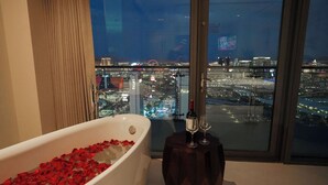 Une salle de bain moderne avec une baignoire remplie de pétales de rose, une grande fenêtre donnant sur la ville et une petite table avec deux verres à vin.