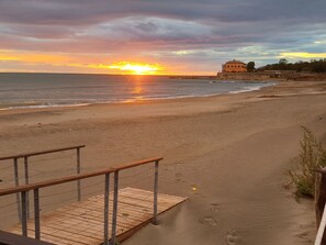 A beach with a wooden walkway leading to a building with a balcony, overlooking the sea and a sunset.