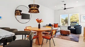 Een moderne woonkamer met een ronde houten eettafel, zwarte en oranje stoelen, een grote spiegel en een plafondventilator.