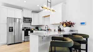 Een moderne keuken met witte kasten, roestvrijstalen apparatuur, een centraal eiland met barstoelen en een decoratieve verlichtingsarmatuur.