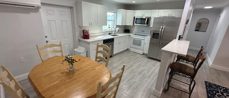 Cocina moderna con gabinetes blancos, un refrigerador de acero inoxidable y un microondas. Hay una mesa de comedor con sillas y un jarrón de flores.