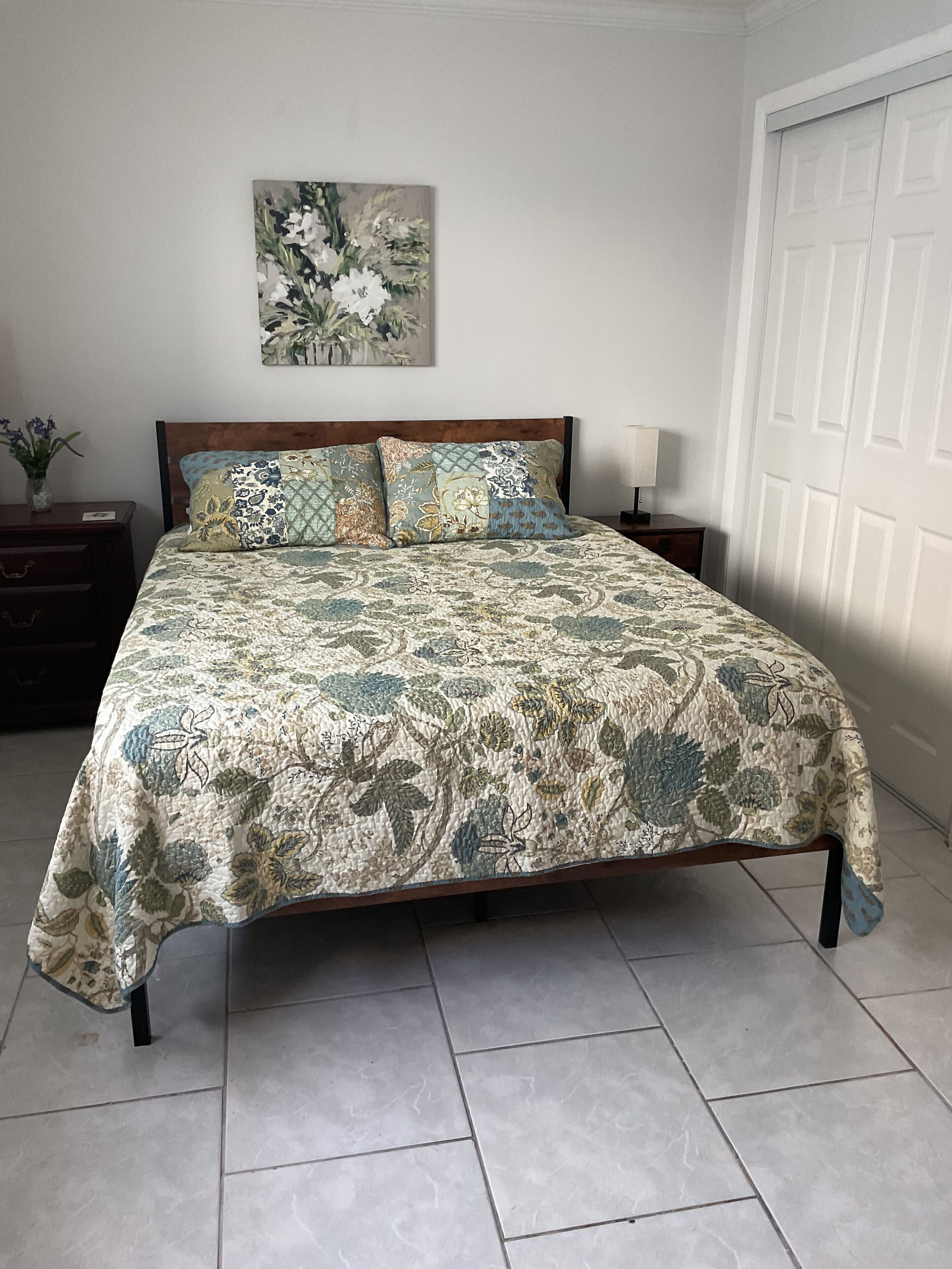 A bedroom with a bed, a nightstand, a lamp, a door, and a wall painting.