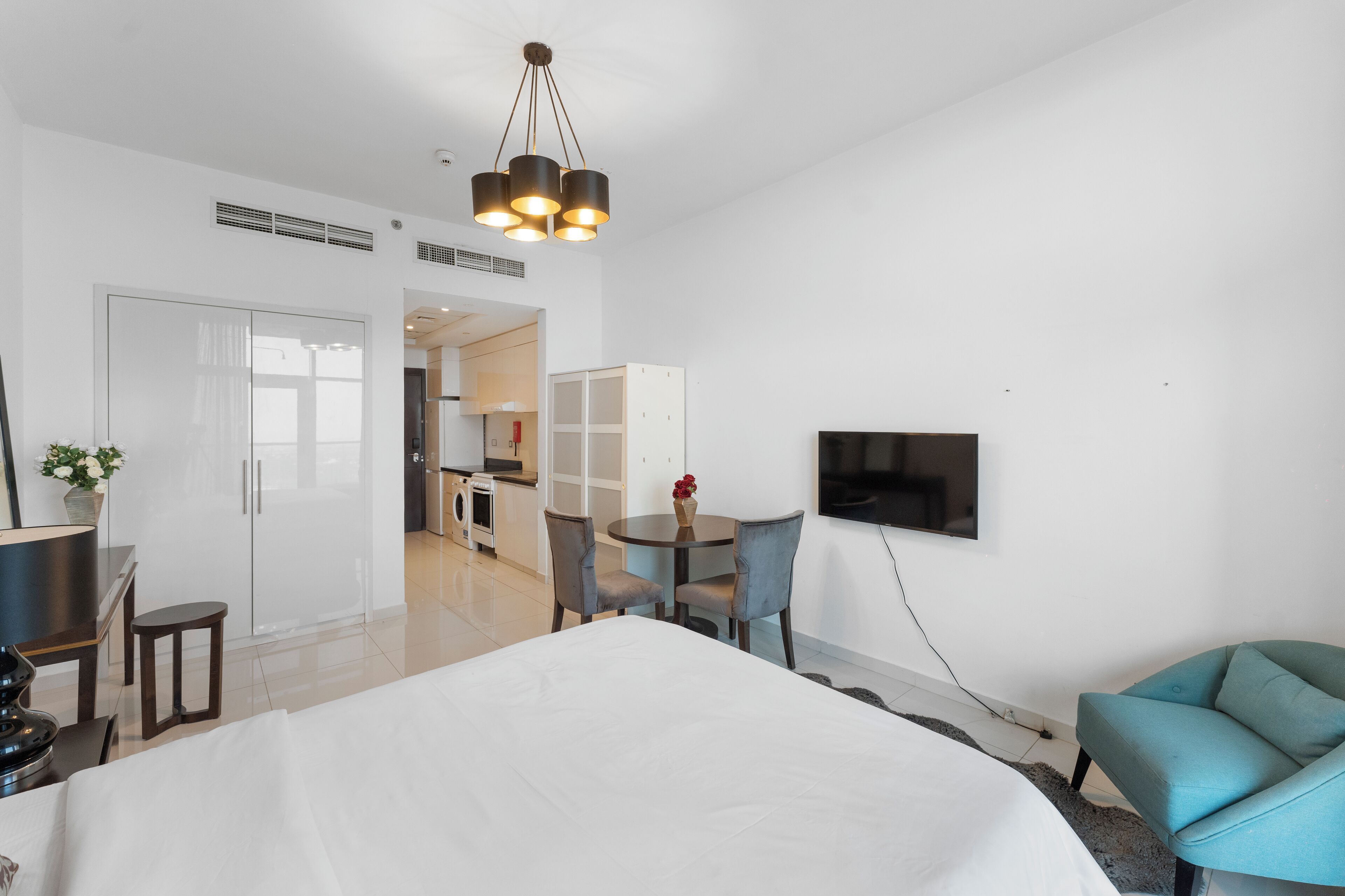 A modern hotel room with a large bed, a dining area with a table and chairs, a TV mounted on the wall, and a kitchenette in the background.
