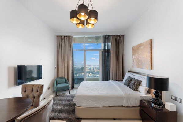 A modern hotel room with a large bed, a TV, a dining area, and a view of the city.