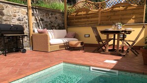 A pool area with a tiled edge, a wicker sofa, a wooden table, and a grill. - Cà La Torre, Stone House with pool in the terrace and super view (Bibola)