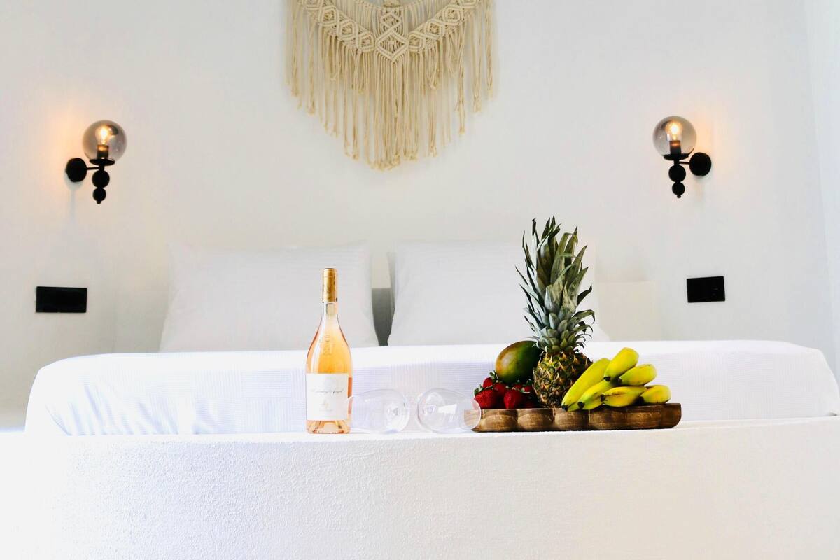 A bedroom with a mural of two people lying on a bed, a macrame wall hanging, a bottle of wine, and a fruit basket.