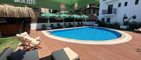 A swimming pool with lounge chairs and umbrellas, surrounded by a wooden deck and a building with balconies.