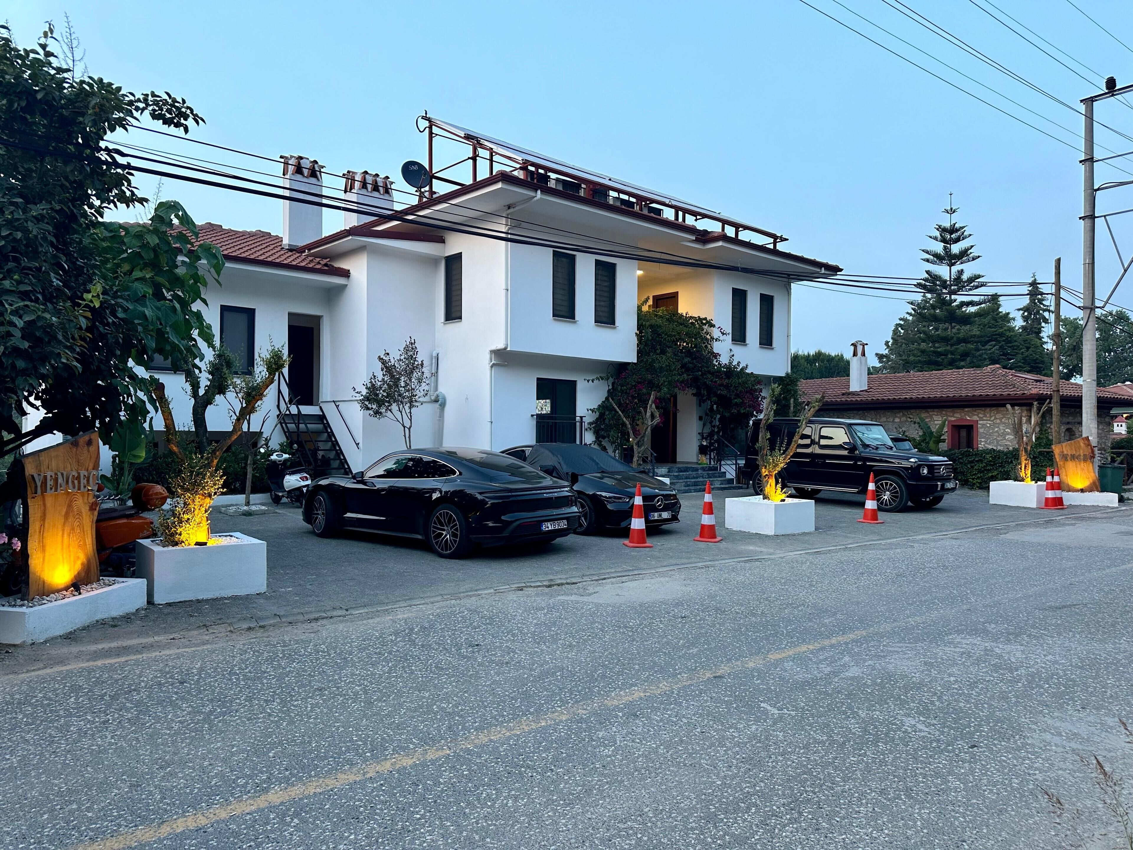 Un edificio moderno de dos pisos con fachada blanca y techo de tejas rojas. Hay dos coches aparcados en la parte delantera, y conos de tráfico naranja están colocados en el suelo.