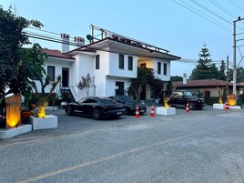 Un edificio moderno de dos pisos con fachada blanca y techo de tejas rojas. Hay dos coches aparcados en la parte delantera, y conos de tráfico naranja están colocados en el suelo.