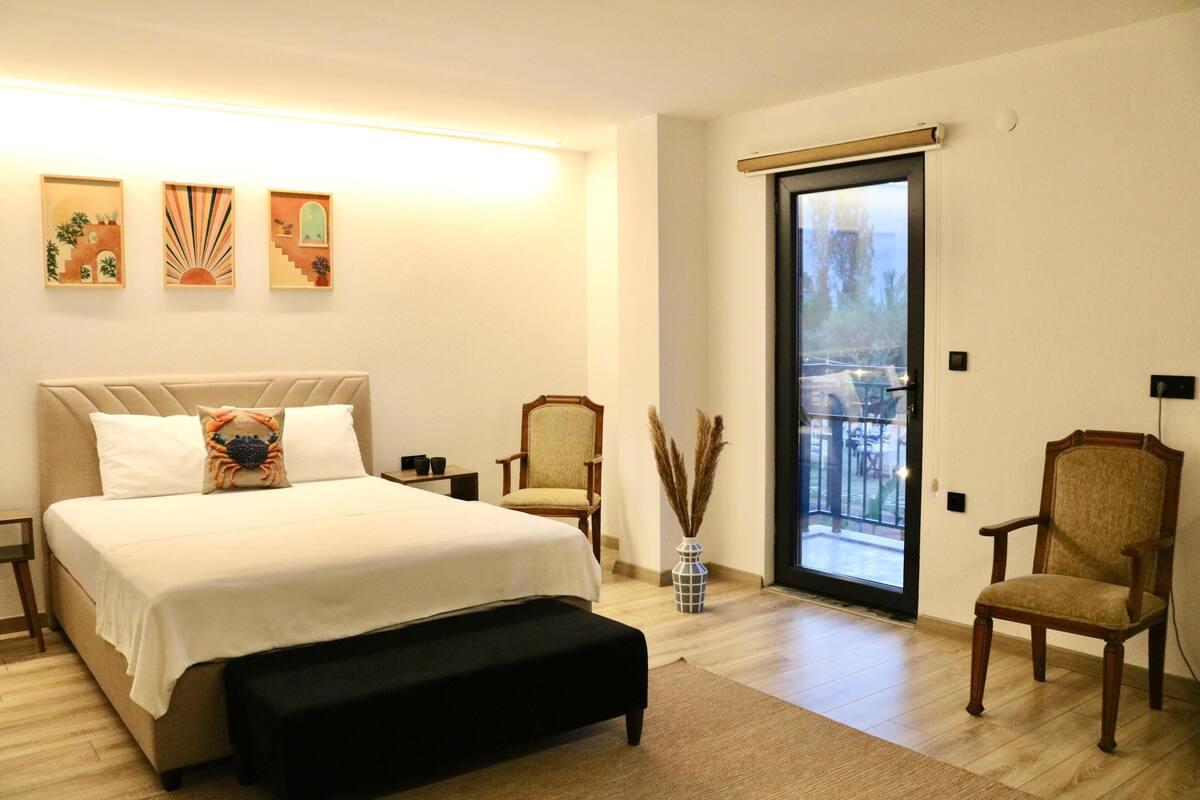 A bedroom with a bed, a bench, a chair, a vase with pampas grass, and a balcony door.