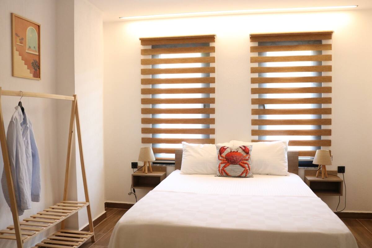 A bedroom with a bed, two nightstands, a wooden drying rack, and two windows with blinds.