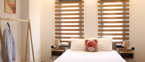 A bedroom with a bed, two nightstands, a wooden drying rack, and two windows with blinds.
