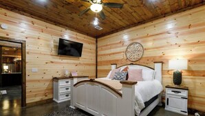A bedroom with a wooden ceiling, a bed with white bedding, a TV mounted on the wall, and a decorative wall piece. - Luxury Cabin | Hot Tub, Fireplace, Game room (Broken Bow)