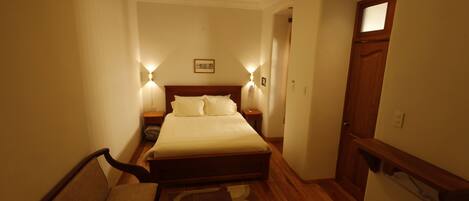 A narrow hotel room with a large bed, two bedside tables, a nightstand, a chair, and a small rug.