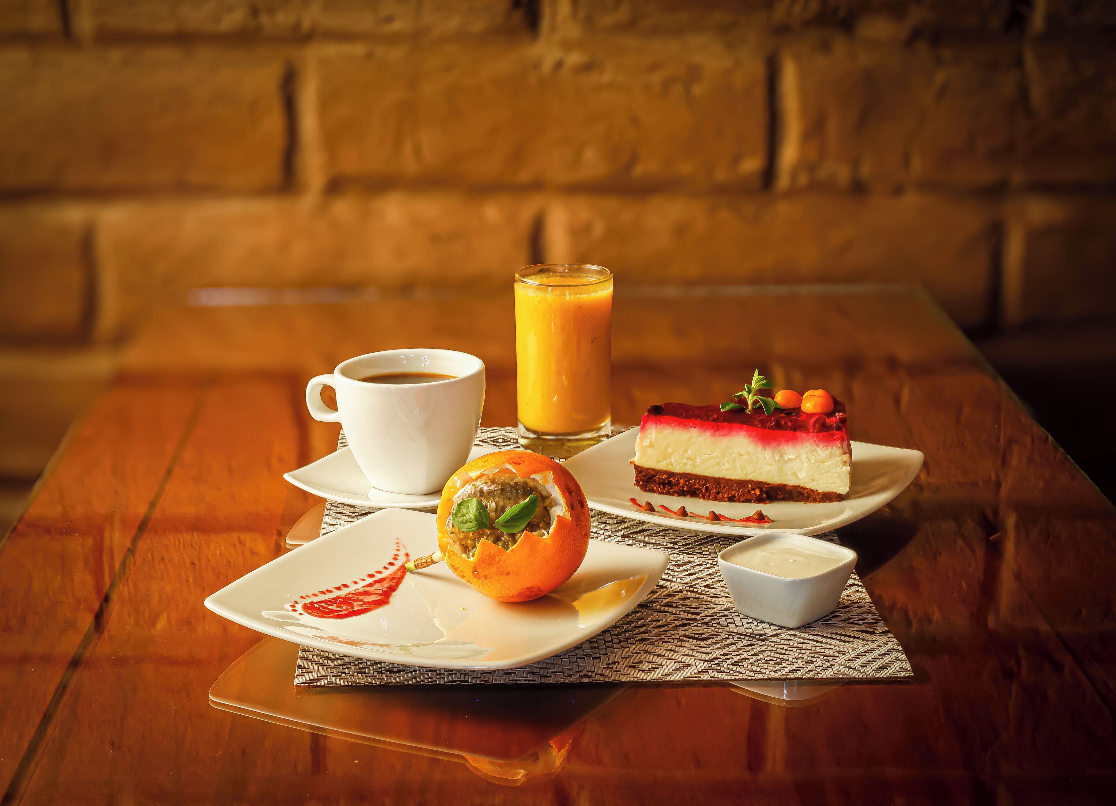 A table with a slice of cheesecake, a cup of coffee, a glass of orange juice, and a small dish of sauce.