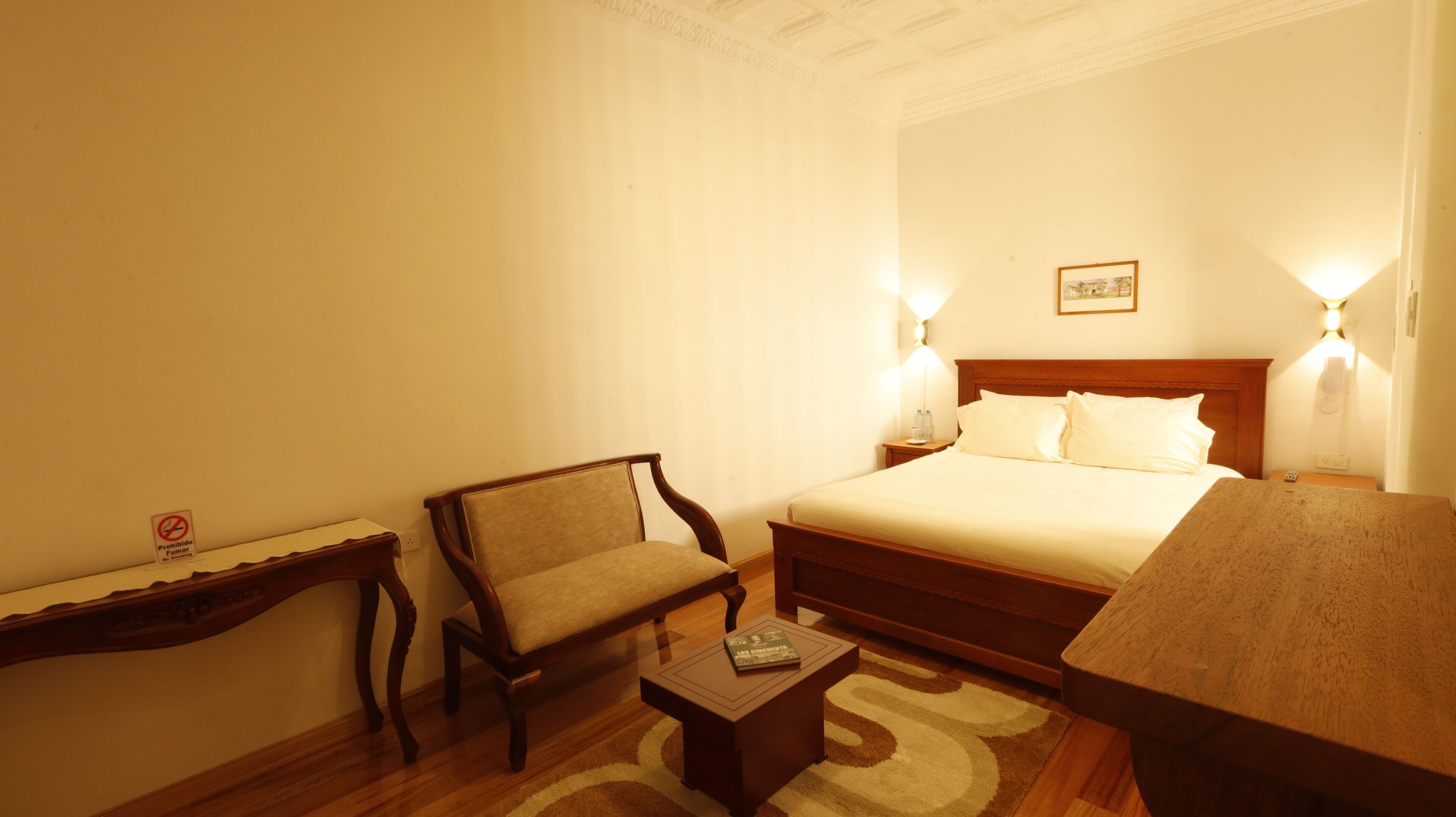 A hotel room with a bed, a wooden chair, a small table, and two wall-mounted lamps.