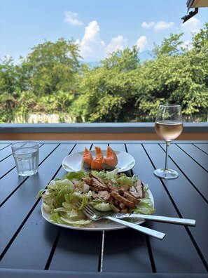 Comida sobre una mesa con una copa de vino blanco y una de agua, al aire libre con árboles al fondo.
