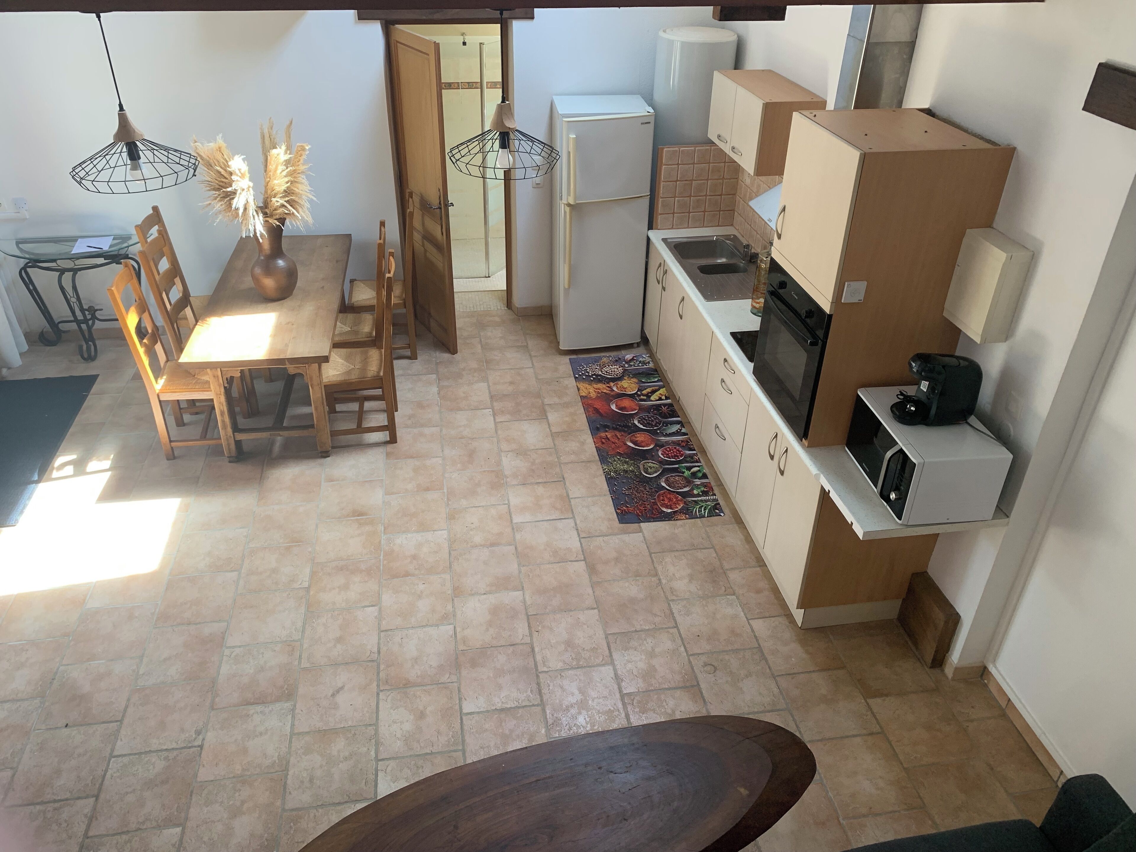 A compact kitchen with a refrigerator, microwave, and stove. There is a dining area with a wooden table and chairs, and a rug with a colorful pattern.