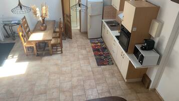 A compact kitchen with a refrigerator, microwave, and stove. There is a dining area with a wooden table and chairs, and a rug with a colorful pattern.
