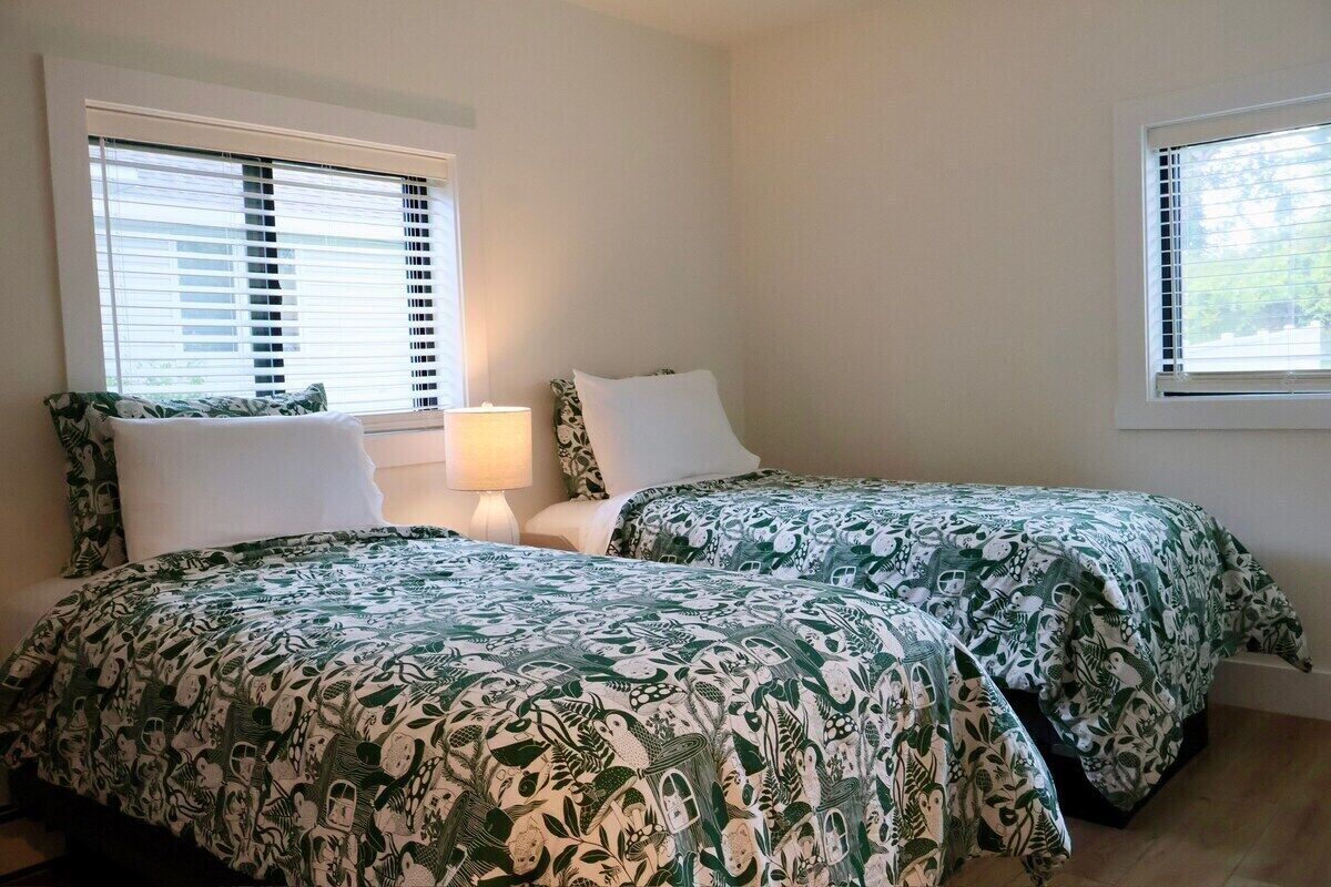 Une chambre avec deux lits, chacun recouvert d’une couvre-lit à motifs verts et blancs, une table de chevet avec une lampe et des fenêtres équipées de stores.