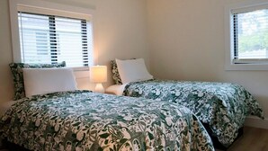 A room with two beds, each with a green and white patterned bedspread, a bedside table with a lamp, and windows with blinds. - Spacious 5 BR House on the River (Indian River)