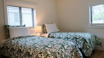 Une chambre avec deux lits, chacun recouvert d’une couvre-lit à motifs verts et blancs, une table de chevet avec une lampe et des fenêtres équipées de stores.