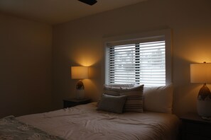 A bedroom with a bed, two bedside lamps, a window with blinds, and a nightstand with a vase. - Spacious 5 BR House on the River (Indian River)