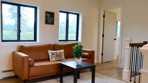 A living room with a brown leather sofa, a black coffee table, and a potted plant. There are two windows providing natural light and a view of greenery outside. - Spacious 5 BR House on the River (Indian River)
