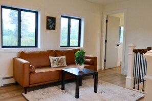 A living room with a brown leather sofa, a black coffee table, and a potted plant. There are two windows providing natural light and a view of greenery outside. - Spacious 5 BR House on the River (Indian River)