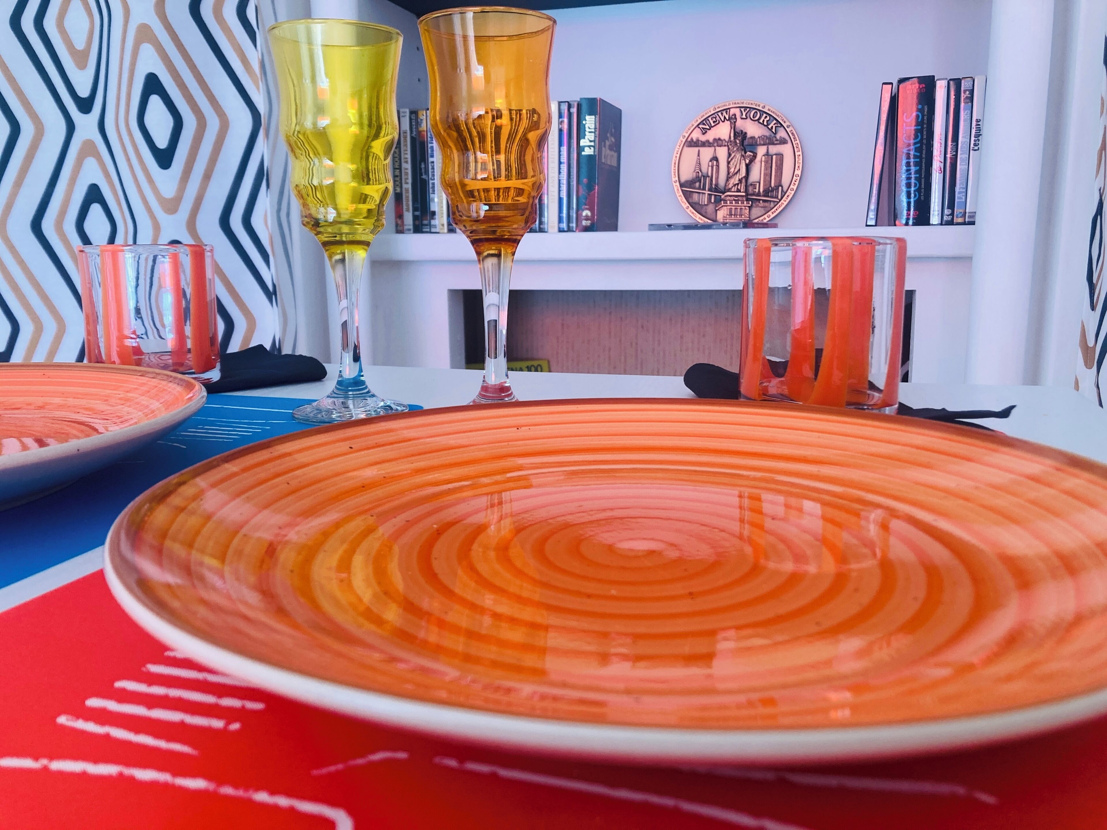 A dining table with orange plates and clear glasses, a patterned wall, and bookshelves with books and a New York City-themed plate.