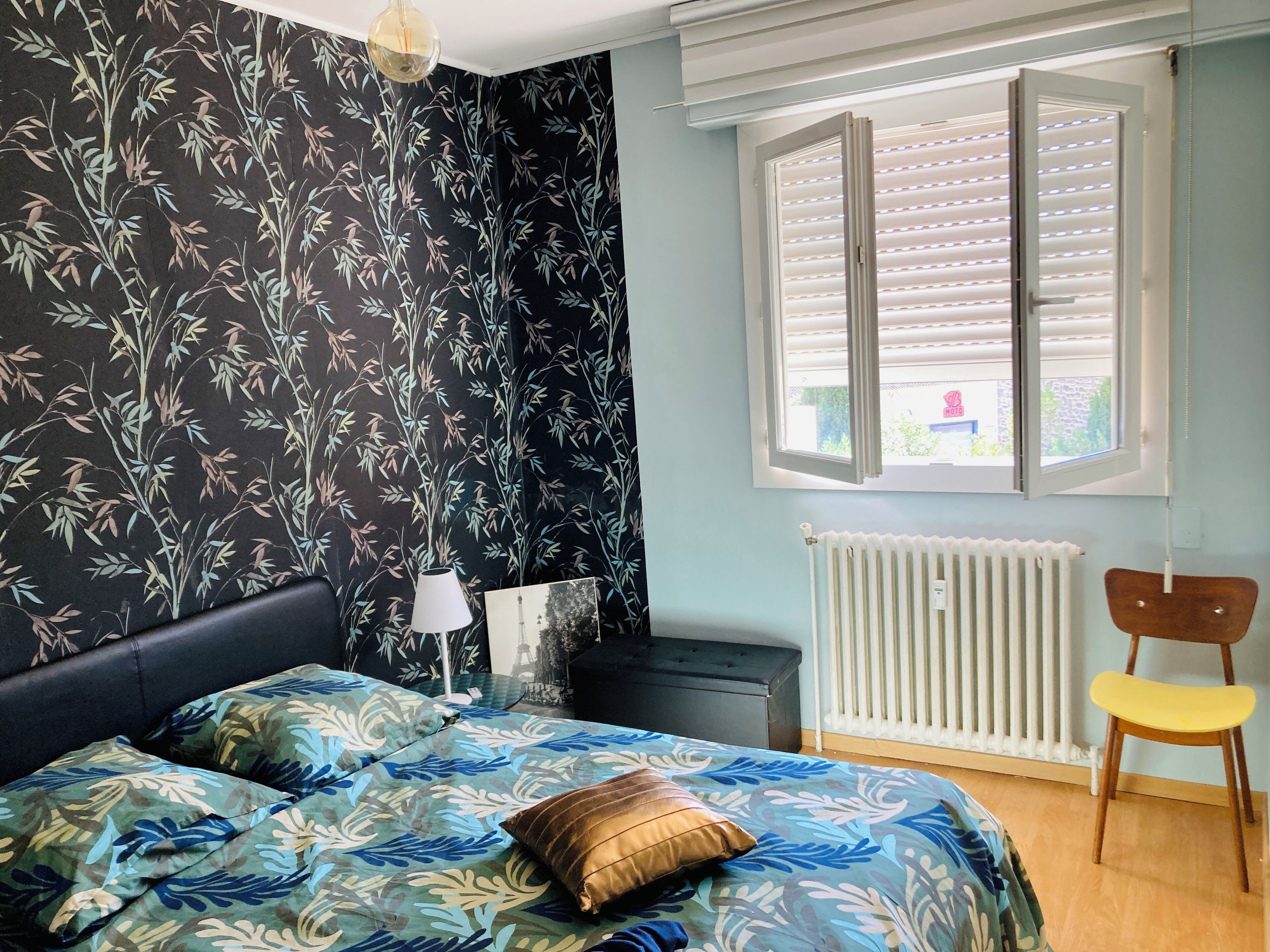 Une chambre à coucher avec un lit, un radiateur, une fenêtre avec des stores, une chaise et une lampe.