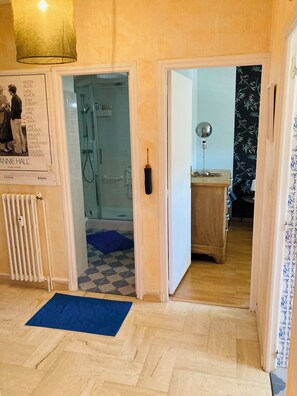 A room with a wooden floor, a blue mat, a radiator, a poster, and a door leading to another room with a bathroom. - Vintage 70' apartment 45 m2 (Castelnau-le-Lez)