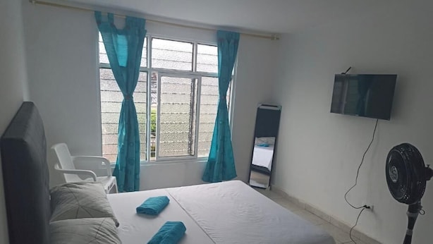 A bedroom with a bed, a TV mounted on the wall, a standing fan, and a window with curtains.