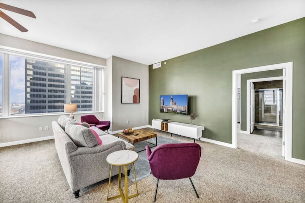 Een moderne woonkamer met een grijze bank, een klein rond tafeltje en een paarse fauteuil. Een flatscreen televisie is aan de muur bevestigd, en een raam biedt uitzicht op de stad.