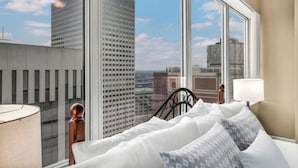 Habitación de hotel con un ventanal grande que ofrece vistas a la ciudad, una cama con ropa de cama blanca y una mesita de noche con una lámpara.