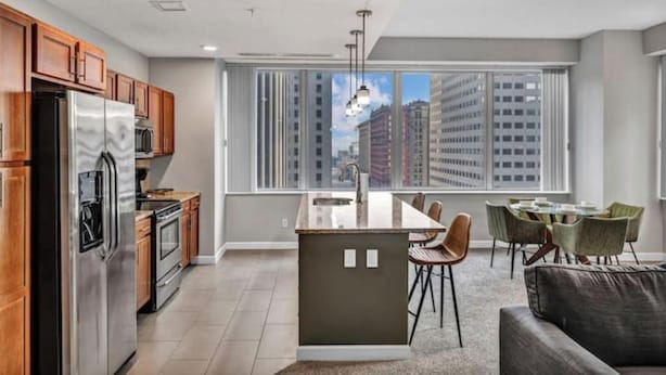 Een moderne keuken met houten kastjes, roestvrijstalen apparatuur en een eethoek met een glazen kookeiland en stoelen.