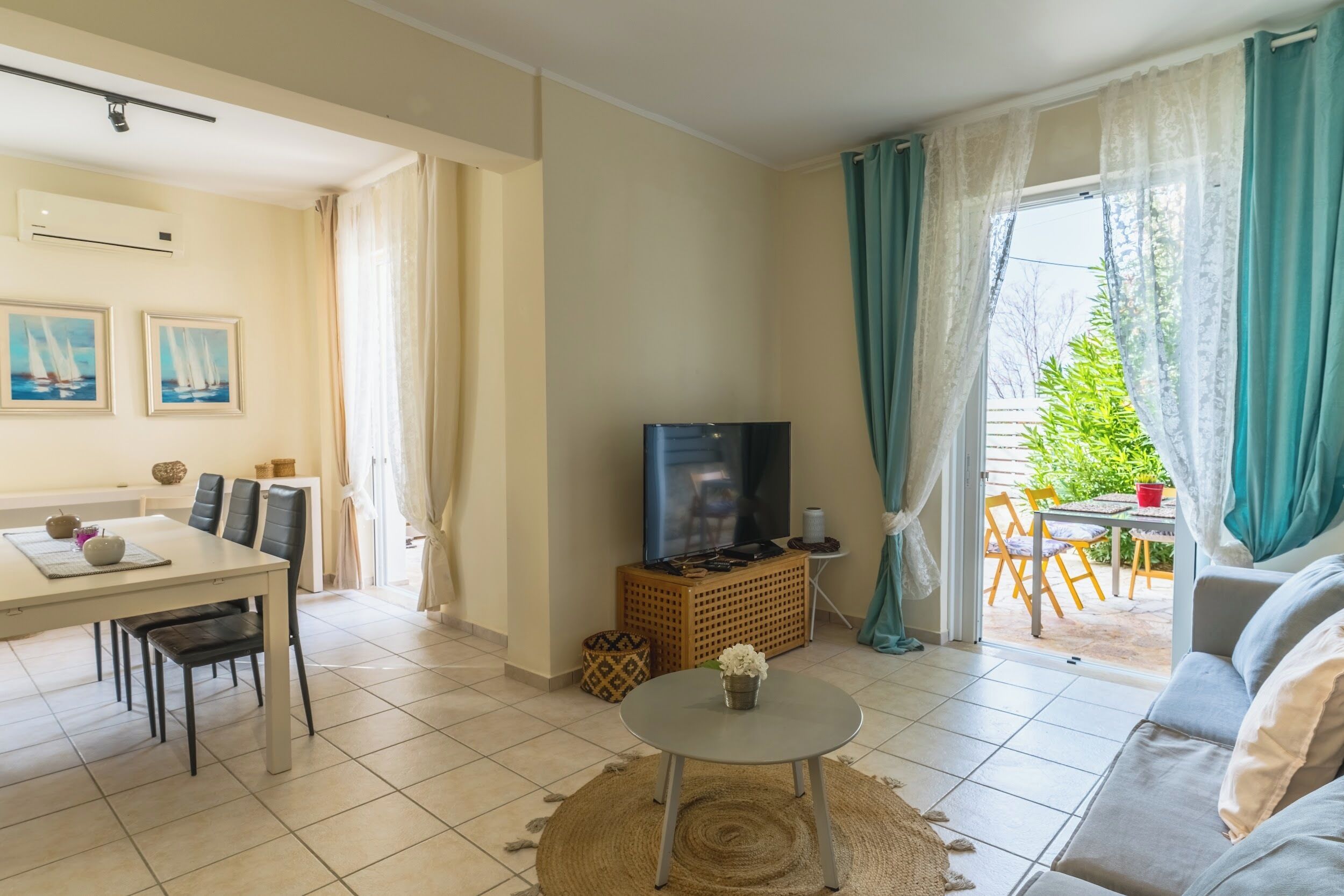 Ein Wohnzimmer mit Fernseher, Esstisch und Stühlen. Ein Sofa und ein kleiner Tisch mit einer Vase sind ebenfalls vorhanden.