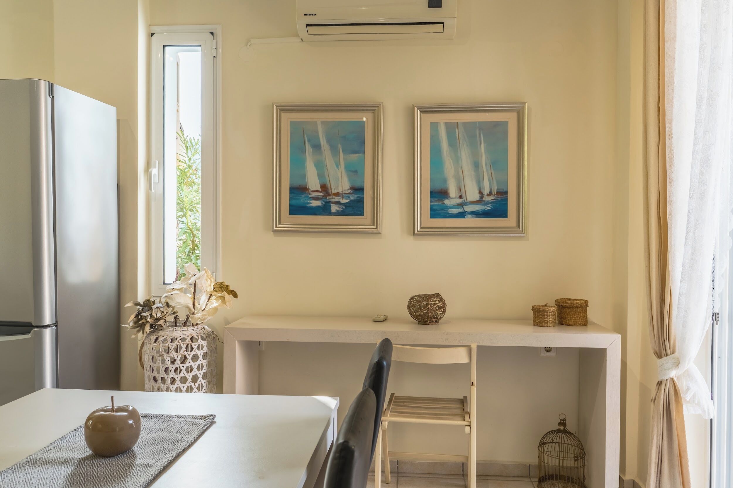 Une cuisine équipée d’un réfrigérateur, d’une table à manger avec un set de table et une pomme, d’une chaise, d’une étagère avec des paniers et de deux tableaux encadrés représentant des voiliers.